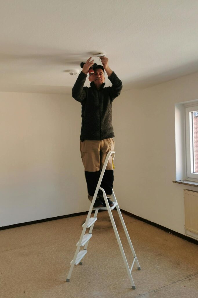 Adult man on ladder fixing a ceiling light in an empty room with beige walls.